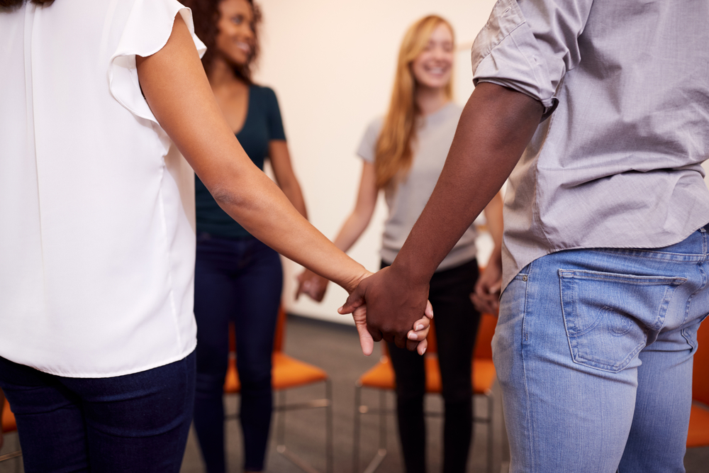 Multi-Cultural Group Of Men And Women Joining Hands At Mental Health Group Therapy Meeting